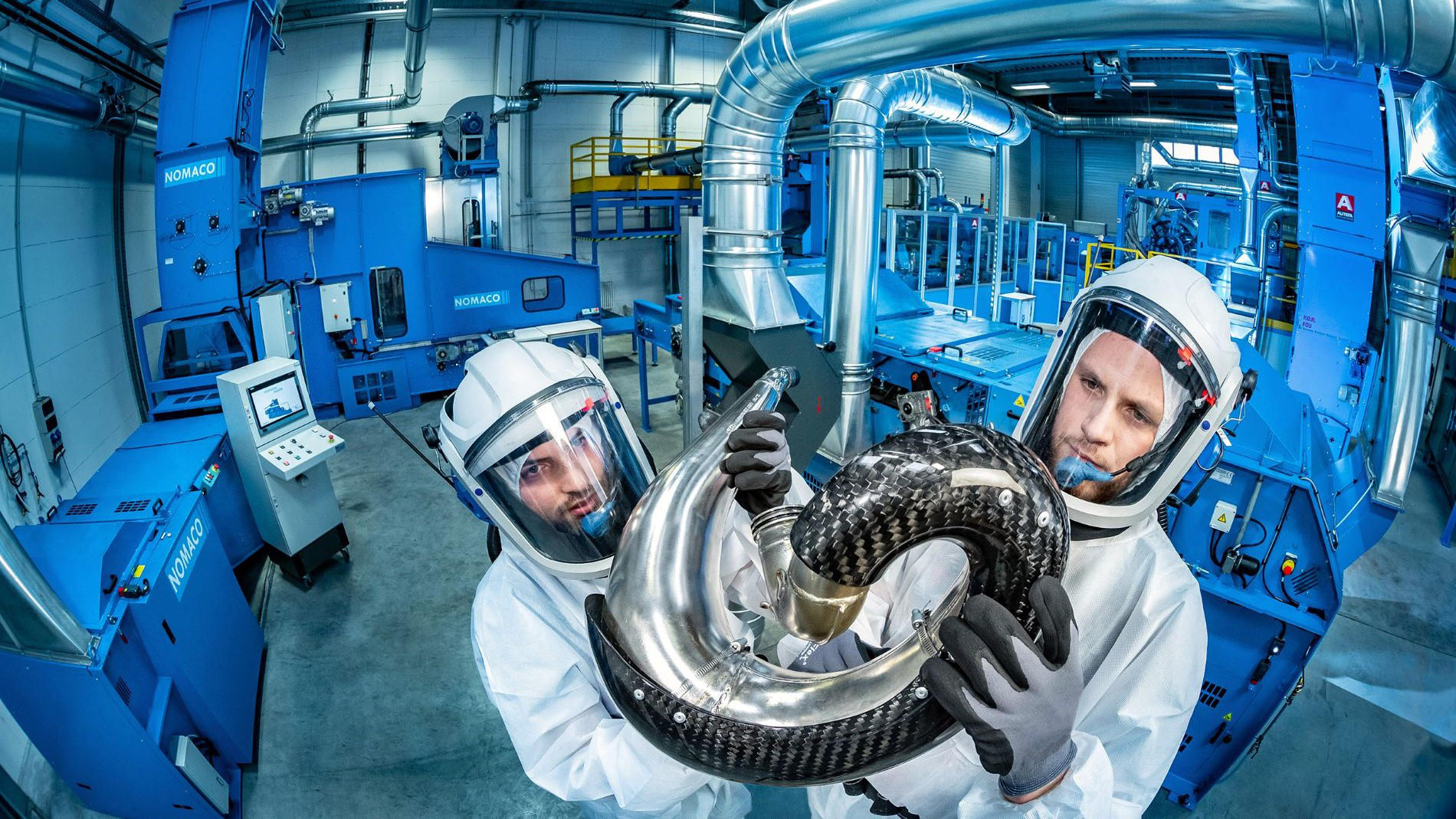 Zwei Fachkräfte in Schutzanzügen halten ein Bauteil aus Carbon in einer modernen Hightech-Anlage zur Werkstoffforschung.