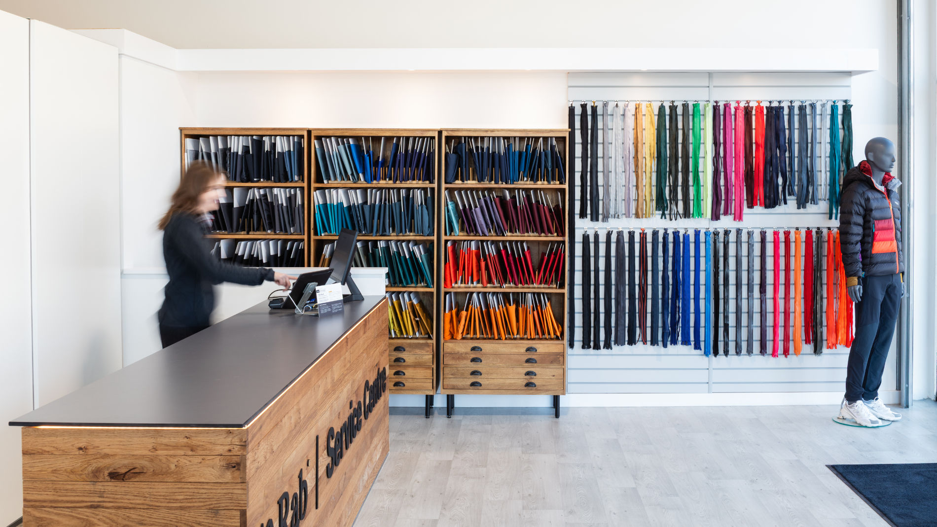 Empfangsbereich eines nachhaltigen Textil-Service-Centers mit Stoffmustern, Reißverschlüssen und Schaufensterpuppe in Outdoor-Bekleidung.
