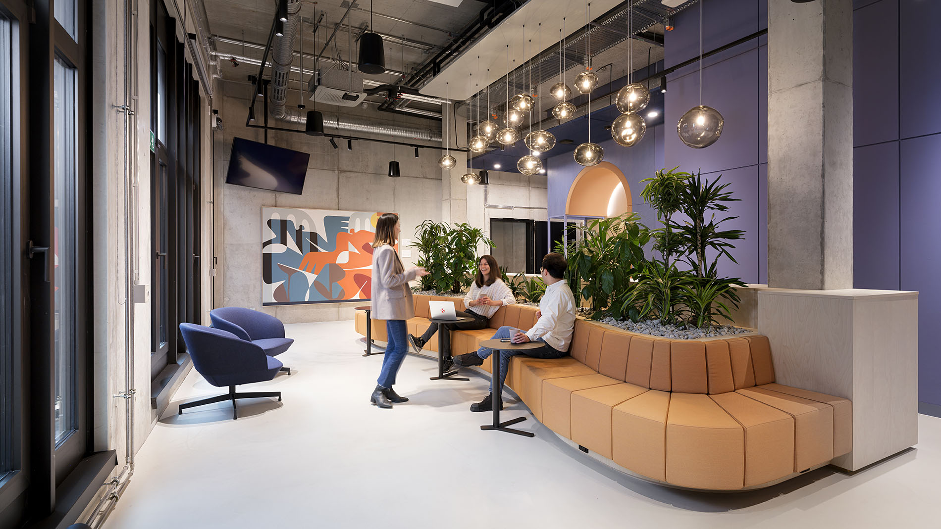Communication zone with curved seating, plant islands, and pendant lights in a modern office setting.