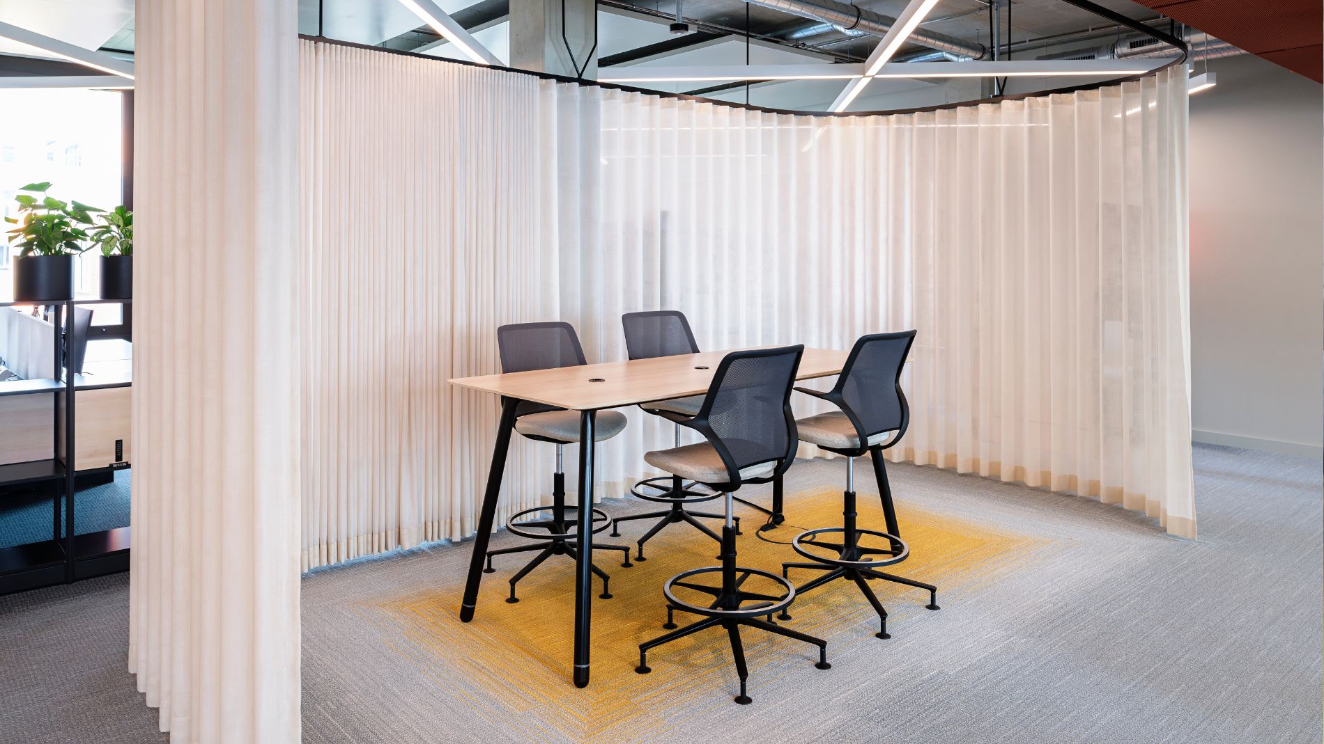 Small meeting area with high chairs and wooden table, surrounded by light curtains for flexible space design.