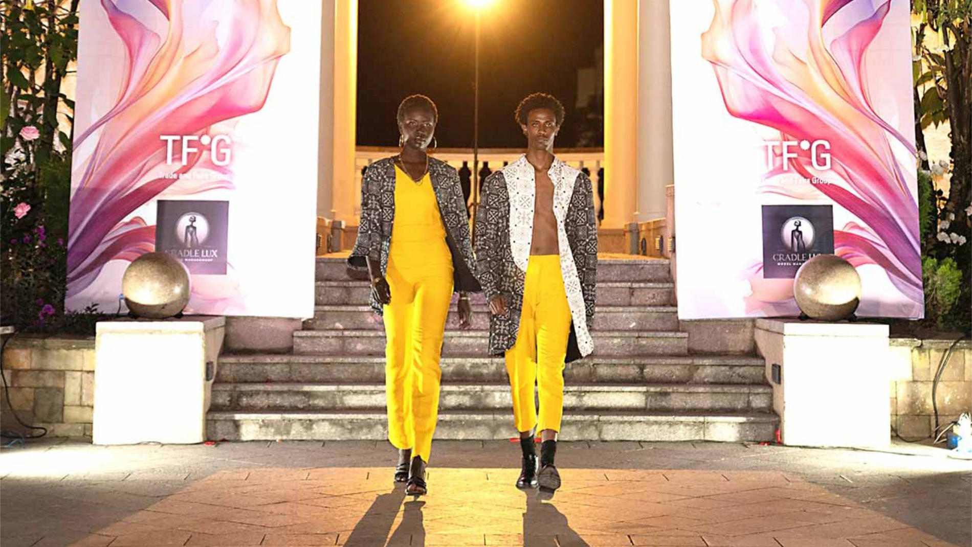 Zwei Models präsentieren auf Outdoor-Laufsteg gelbe Outfits mit gemusterten Mänteln bei einer Modenschau
